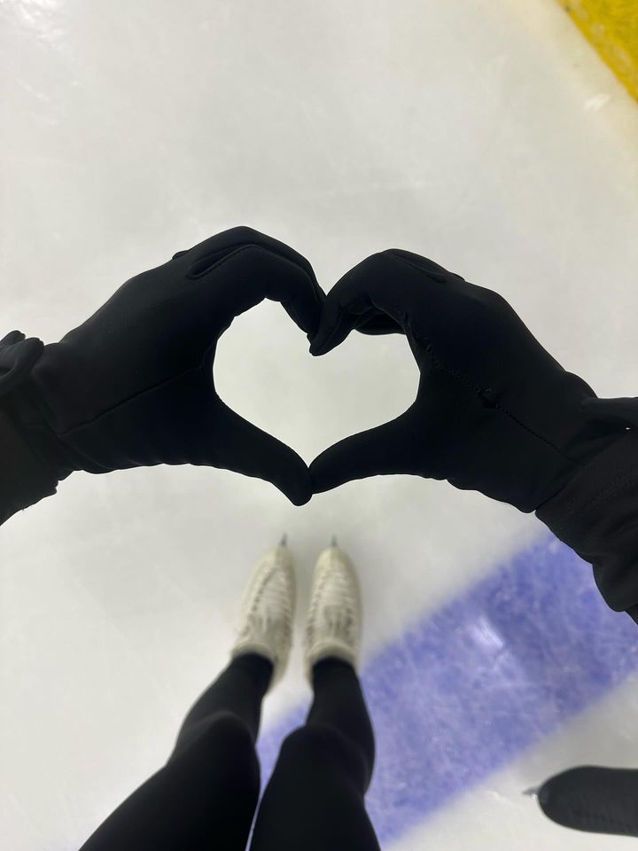 A first-person view of someone on an ice rink wearing black gloves and black leggings, forming a heart shape with their hands. Their white ice skates are visible below, with the ice surface and a blue rink marking in the background.