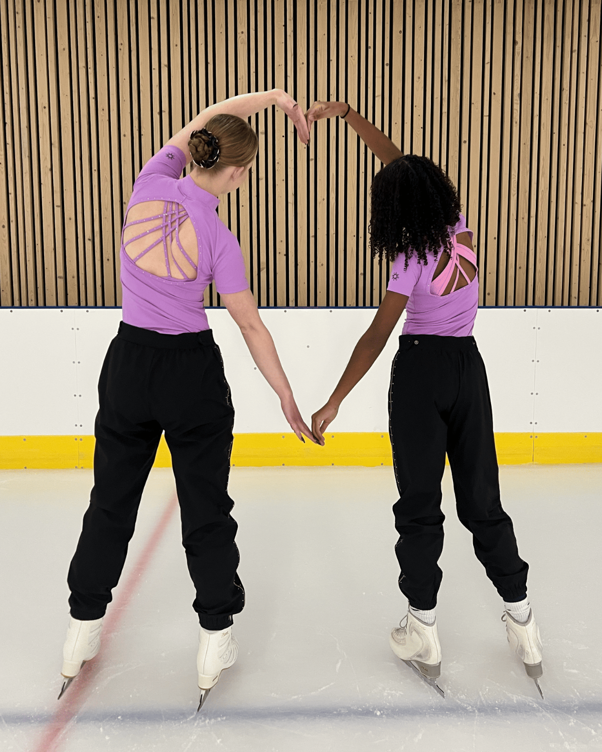 Two ice skaters pose on an indoor ice rink, wearing matching purple athletic tops with a cutout back design and black pants, holding hands to form a heart shape with their raised arms.